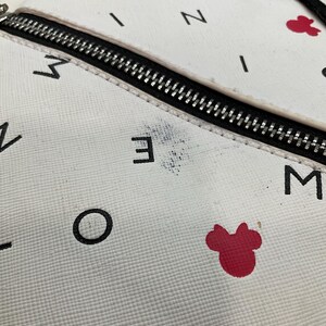 May include: White backpack with a black zipper and black lettering. The backpack has red Minnie Mouse bow accents and red Mickey Mouse head accents. The backpack has a textured surface.