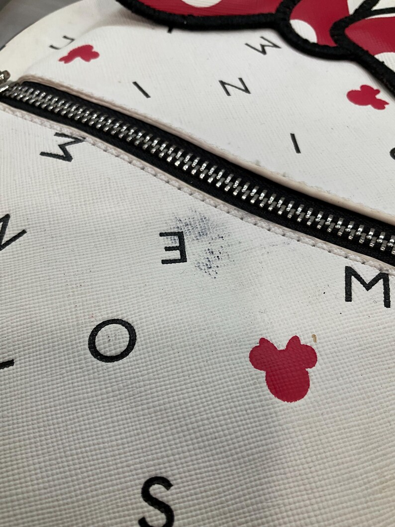 May include: White backpack with a black zipper and black lettering. The backpack has red Minnie Mouse bow accents and red Mickey Mouse head accents. The backpack has a textured surface.