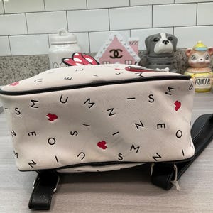 May include: A white backpack with black trim, featuring a pattern of letters, red hearts, and the letter "M". The bag has black shoulder straps and a top handle. The design is likely inspired by a popular cartoon character.