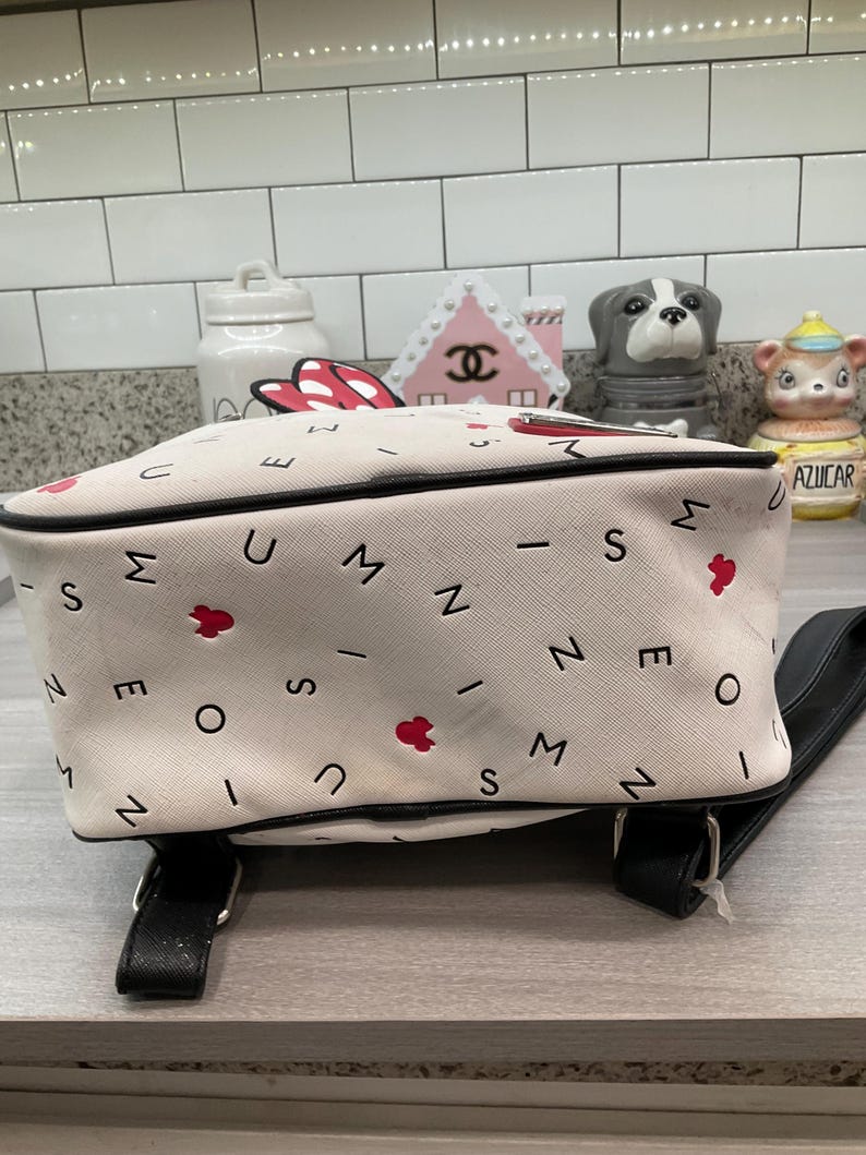 May include: A white backpack with black trim, featuring a pattern of letters, red hearts, and the letter "M". The bag has black shoulder straps and a top handle. The design is likely inspired by a popular cartoon character.