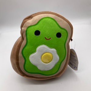 Puede incluir: Un juguete de peluche de tostada de aguacate con una cara sonriente, relleno de aguacate verde y un huevo frito blanco con yema amarilla. El juguete es marrón en los bordes para parecerse a pan.