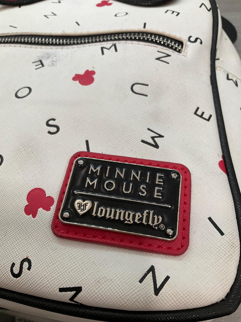 May include: White backpack with a pattern of black letters and red Minnie Mouse silhouettes. A rectangular black and red patch reads "MINNIE MOUSE" and "loungefly" in silver lettering.