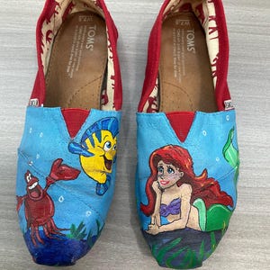 May include: A pair of TOMS slip-on shoes, size W7.5, with hand-painted illustrations of characters from The Little Mermaid. The shoes have a red trim and a blue canvas upper with Ariel, Flounder, and Sebastian.