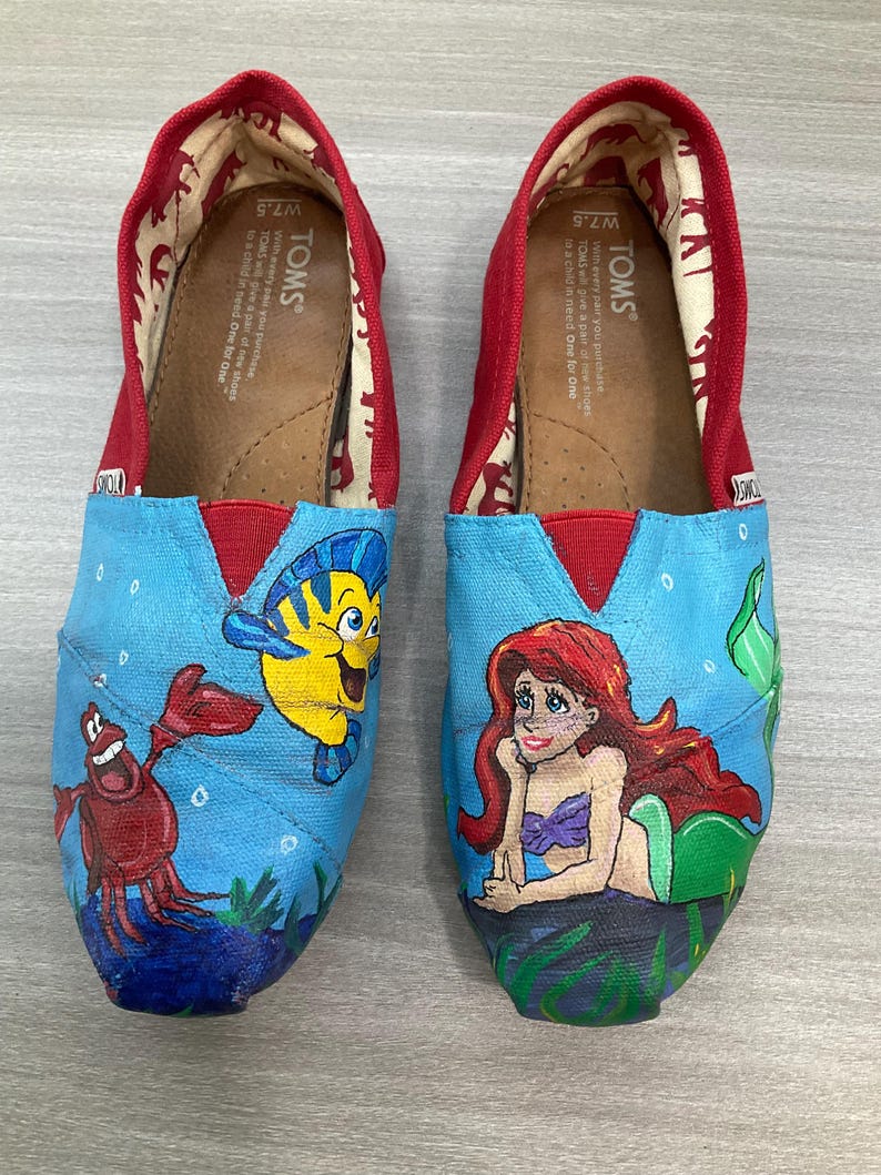 May include: A pair of TOMS slip-on shoes, size W7.5, with hand-painted illustrations of characters from The Little Mermaid. The shoes have a red trim and a blue canvas upper with Ariel, Flounder, and Sebastian.