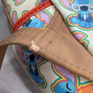 May include: Cream-colored bag featuring a repeating pattern of the cartoon character Stitch in various poses. The bag has a brown strap and red trim. The Stitch illustrations are outlined in rainbow colors. The bag appears to be made of a leather-like material.