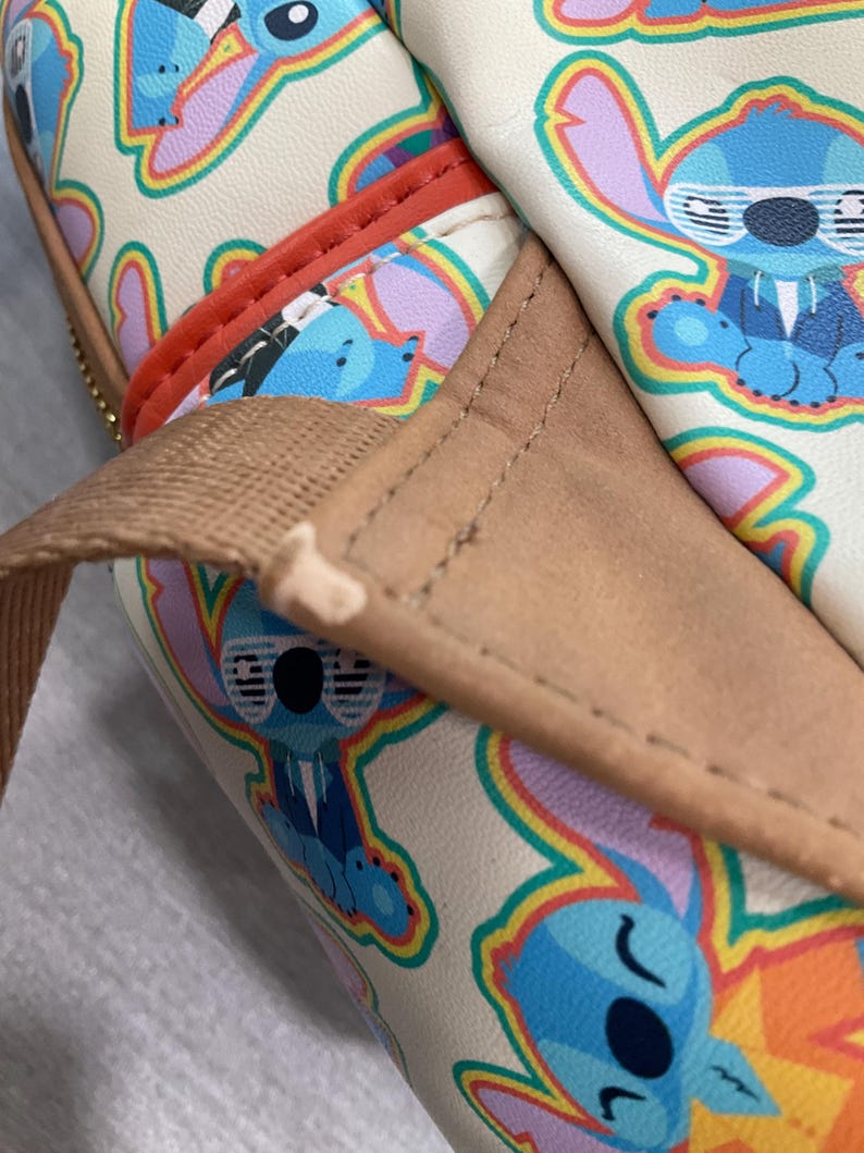 May include: Cream-colored bag featuring a repeating pattern of the cartoon character Stitch in various poses. The bag has a brown strap and red trim. The Stitch illustrations are outlined in rainbow colors. The bag appears to be made of a leather-like material.