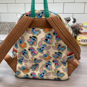 May include: A beige backpack featuring a repeating pattern of the character Stitch in various outfits. The backpack has brown straps and trim, a green handle, and red accents. The bag is sitting on a light gray surface.