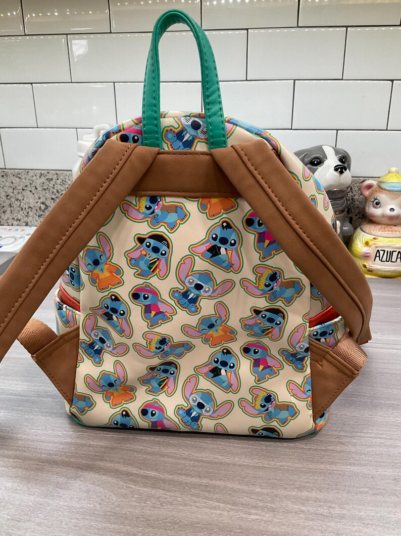 May include: A beige backpack featuring a repeating pattern of the character Stitch in various outfits. The backpack has brown straps and trim, a green handle, and red accents. The bag is sitting on a light gray surface.