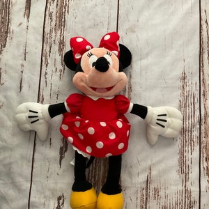 May include: A Minnie Mouse plush toy. The doll has a red dress with white polka dots, a matching bow, and yellow shoes. The doll has black ears, arms, and legs, and white gloves. The plush toy is lying on a light-colored wooden surface.