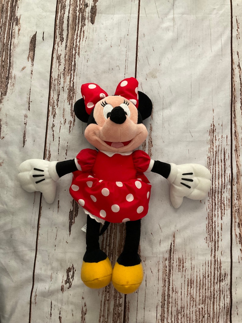 May include: A Minnie Mouse plush toy. The doll has a red dress with white polka dots, a matching bow, and yellow shoes. The doll has black ears, arms, and legs, and white gloves. The plush toy is lying on a light-colored wooden surface.