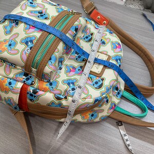 May include: A cream-colored backpack featuring a repeating pattern of the character Stitch in various poses. The bag has brown trim, a teal zipper, and adjustable straps. The bag measures approximately 10 inches in height and 8 inches in width.