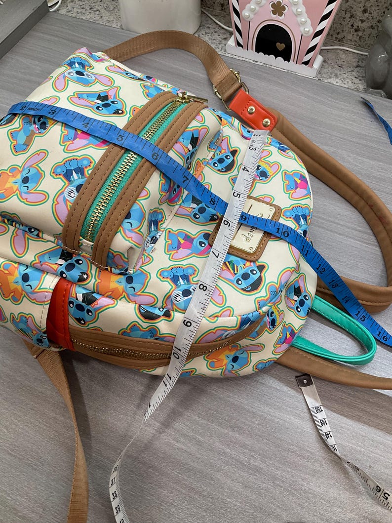 May include: A cream-colored backpack featuring a repeating pattern of the character Stitch in various poses. The bag has brown trim, a teal zipper, and adjustable straps. The bag measures approximately 10 inches in height and 8 inches in width.