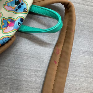 May include: A cream-colored backpack featuring a repeating pattern of the cartoon character Stitch in various poses. The bag has brown straps and a teal handle. The bag is made of a smooth material.