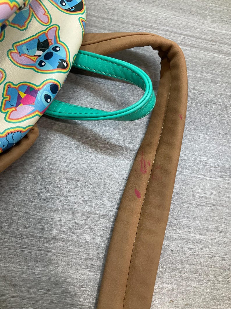 May include: A cream-colored backpack featuring a repeating pattern of the cartoon character Stitch in various poses. The bag has brown straps and a teal handle. The bag is made of a smooth material.