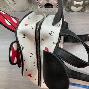 May include: A white backpack with black accents, designed to resemble a cartoon character. The bag features a zipper closure, black handles, and a red and white polka dot bow. The bag is decorated with red hearts and the letters "M", "I", "N", "O", and "S".