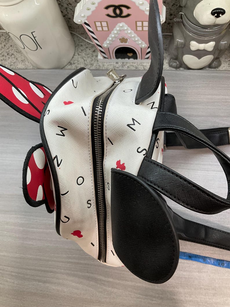 May include: A white backpack with black accents, designed to resemble a cartoon character. The bag features a zipper closure, black handles, and a red and white polka dot bow. The bag is decorated with red hearts and the letters "M", "I", "N", "O", and "S".
