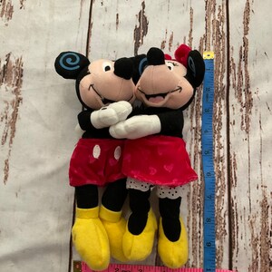 Puede incluir: Una pareja de peluche de Mickey Mouse y Minnie Mouse abraz&aacute;ndose. Mickey lleva pantalones cortos rojos y Minnie lleva una falda roja con lunares blancos. Ambos llevan zapatos amarillos.