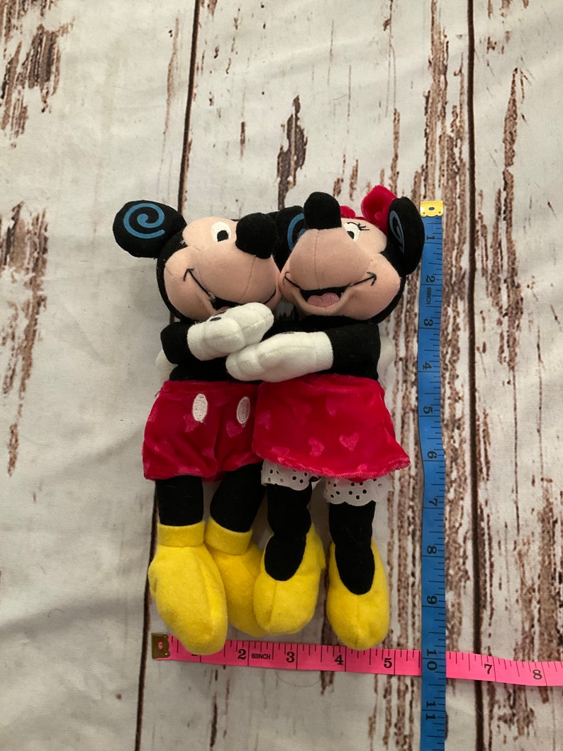 Puede incluir: Una pareja de peluche de Mickey Mouse y Minnie Mouse abraz&aacute;ndose. Mickey lleva pantalones cortos rojos y Minnie lleva una falda roja con lunares blancos. Ambos llevan zapatos amarillos.
