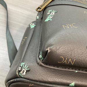 May include: A dark gray backpack with a repeating pattern of Mickey Mouse dressed as the Statue of Liberty and the letters "NYC" in gold. The backpack has a black strap and a gold zipper.