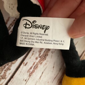 May include: A close-up of a Disney product tag. The tag is white with black text, including the Disney logo and copyright information. The tag is attached to a black plush toy with a red and white accent.