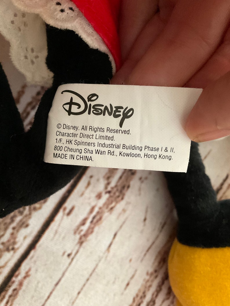 May include: A close-up of a Disney product tag. The tag is white with black text, including the Disney logo and copyright information. The tag is attached to a black plush toy with a red and white accent.