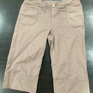 May include: Tan capri pants with vertical pinstripes. The pants have two front pockets, a button closure, and wide cuffs at the hem. The fabric appears to be a lightweight material, suitable for warmer weather.