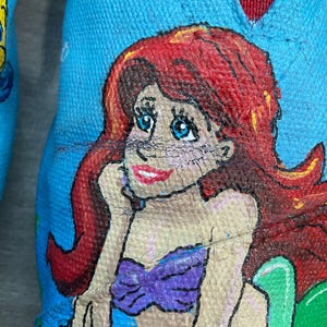 May include: Close-up of a blue shoe with a hand-painted illustration of a mermaid. The mermaid has red hair, blue eyes, and a purple top. The shoe's interior is red.