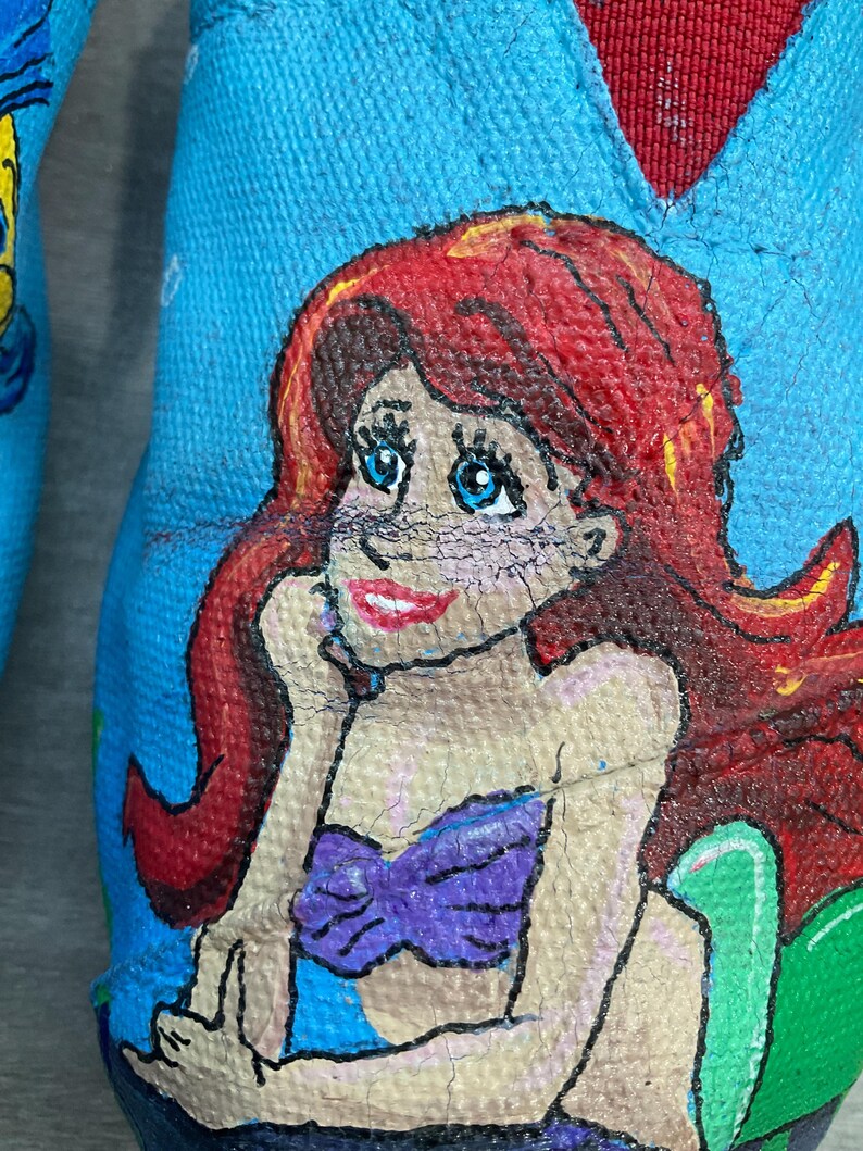 May include: Close-up of a blue shoe with a hand-painted illustration of a mermaid. The mermaid has red hair, blue eyes, and a purple top. The shoe's interior is red.