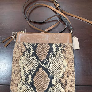 May include: A brown leather crossbody bag with a snakeskin print on the lower half. The bag has a long adjustable strap and a small tag. The top portion of the bag is a solid brown color, and the bag has a zipper closure.