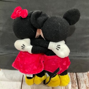 Puede incluir: Un peluche de Mickey Mouse y Minnie Mouse abraz&aacute;ndose. Mickey lleva pantalones cortos rojos y Minnie lleva un vestido rojo con un lazo rojo. Ambos tienen orejas negras y zapatos amarillos.