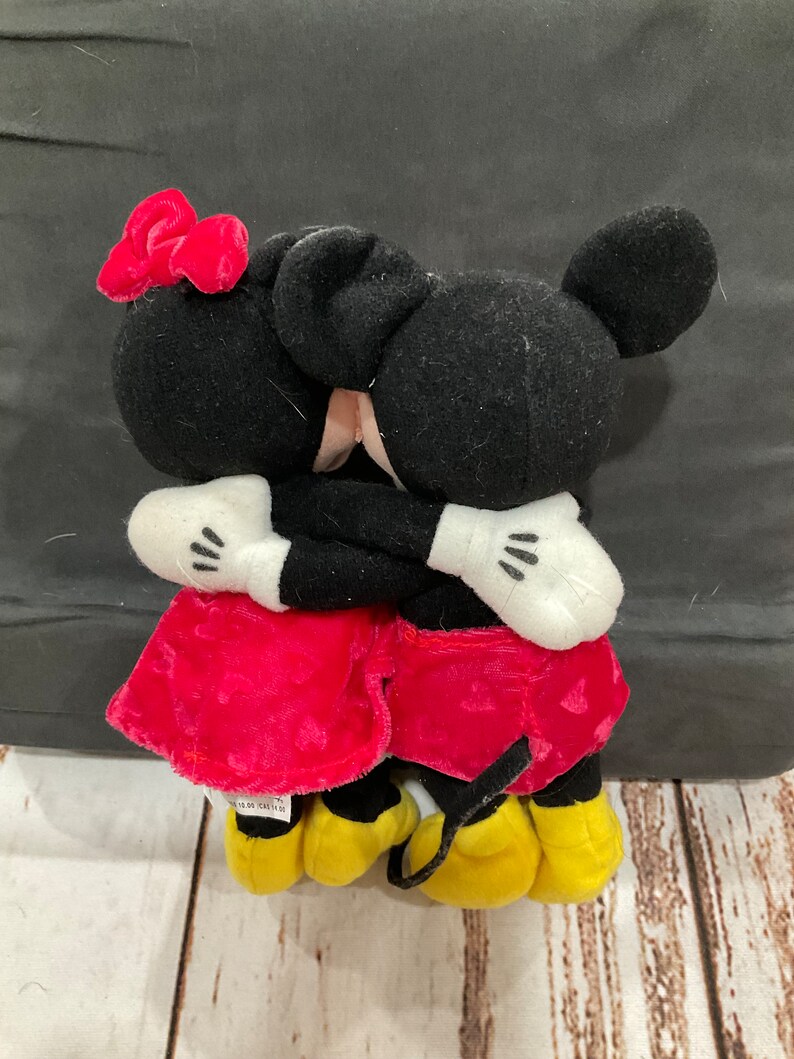 Puede incluir: Un peluche de Mickey Mouse y Minnie Mouse abraz&aacute;ndose. Mickey lleva pantalones cortos rojos y Minnie lleva un vestido rojo con un lazo rojo. Ambos tienen orejas negras y zapatos amarillos.