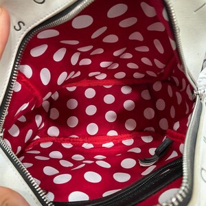 May include: White handbag with a black zipper and a red interior with white polka dots. The bag has black trim and the letters "S" and "O" are visible on the exterior.