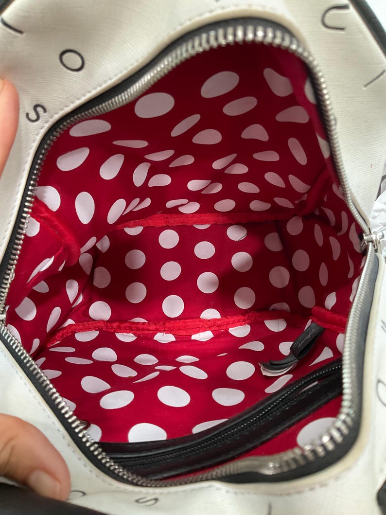 May include: White handbag with a black zipper and a red interior with white polka dots. The bag has black trim and the letters "S" and "O" are visible on the exterior.