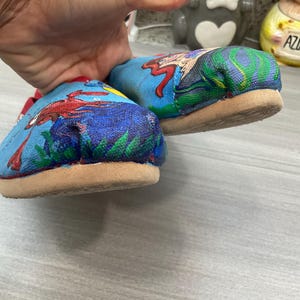May include: A pair of blue canvas shoes with a colourful illustration of a crab and a mermaid. The shoes have a tan sole and red trim. The artwork is painted on the top of the shoes.