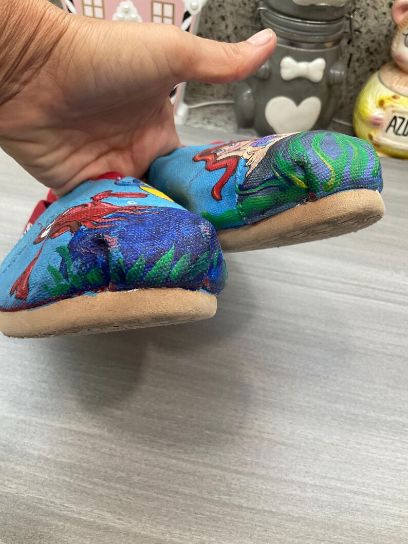 May include: A pair of blue canvas shoes with a colourful illustration of a crab and a mermaid. The shoes have a tan sole and red trim. The artwork is painted on the top of the shoes.
