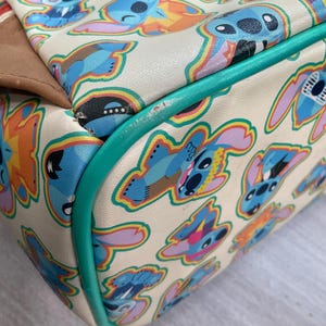 May include: Cream-colored handbag featuring a repeating pattern of blue cartoon character illustrations with colorful accents. The bag has a teal green trim and a brown accent on the side.