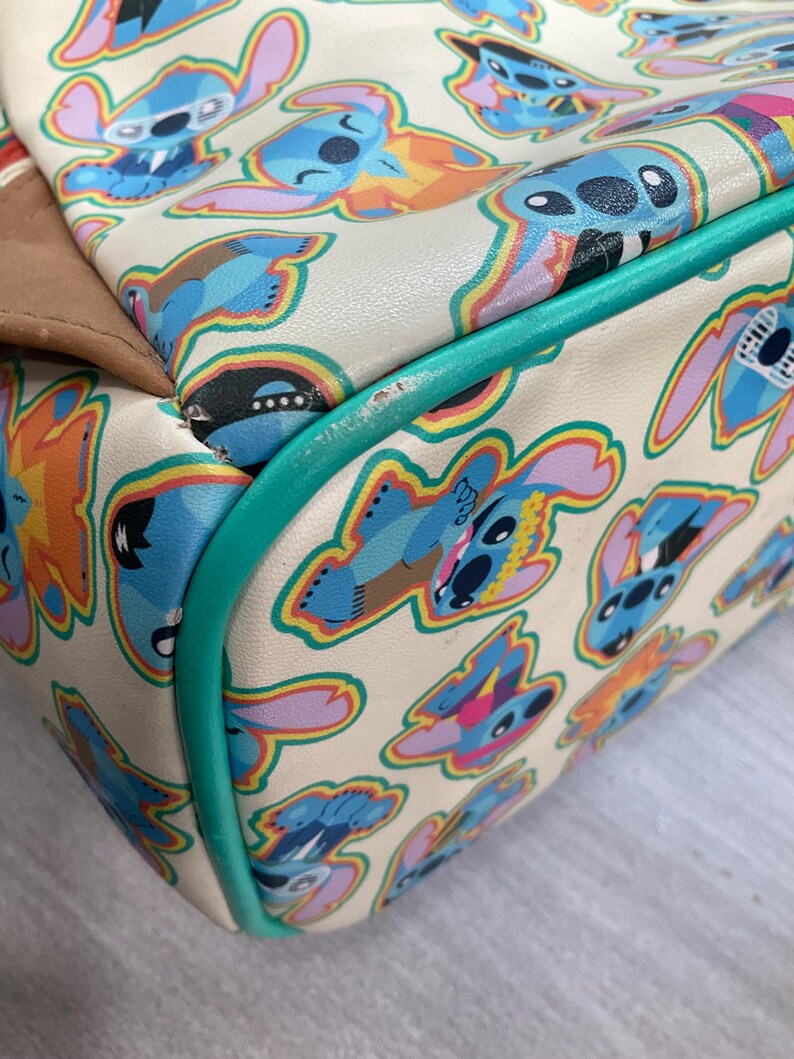 May include: Cream-colored handbag featuring a repeating pattern of blue cartoon character illustrations with colorful accents. The bag has a teal green trim and a brown accent on the side.