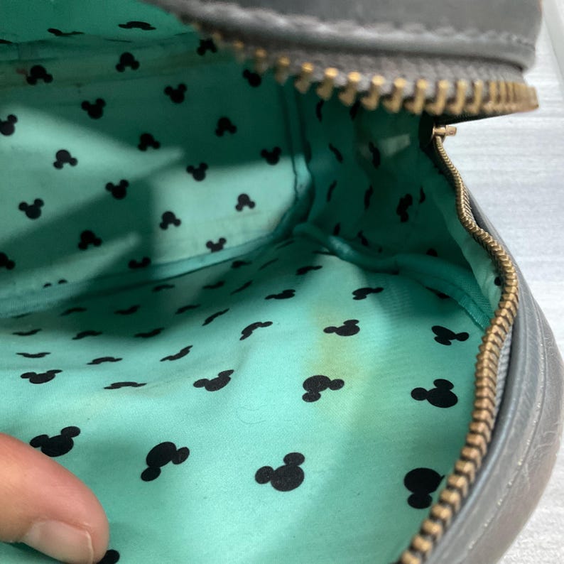 May include: Gray zippered bag with a teal interior patterned with black Mickey Mouse silhouettes. The bag's interior shows some discoloration. The zipper is a brass color.