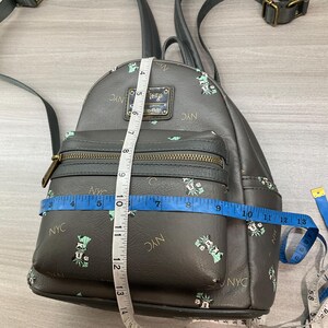 May include: A black faux leather backpack with a front zippered pocket. The backpack features a pattern of Mickey Mouse dressed as the Statue of Liberty and the text "NYC". The backpack measures approximately 10 inches wide.