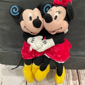 Puede incluir: Una pareja de peluche de Mickey Mouse y Minnie Mouse tomados de la mano. Mickey lleva pantalones cortos rojos y zapatos amarillos. Minnie lleva un vestido rojo con lunares blancos y zapatos amarillos. Ambos est&aacute;n sonriendo.