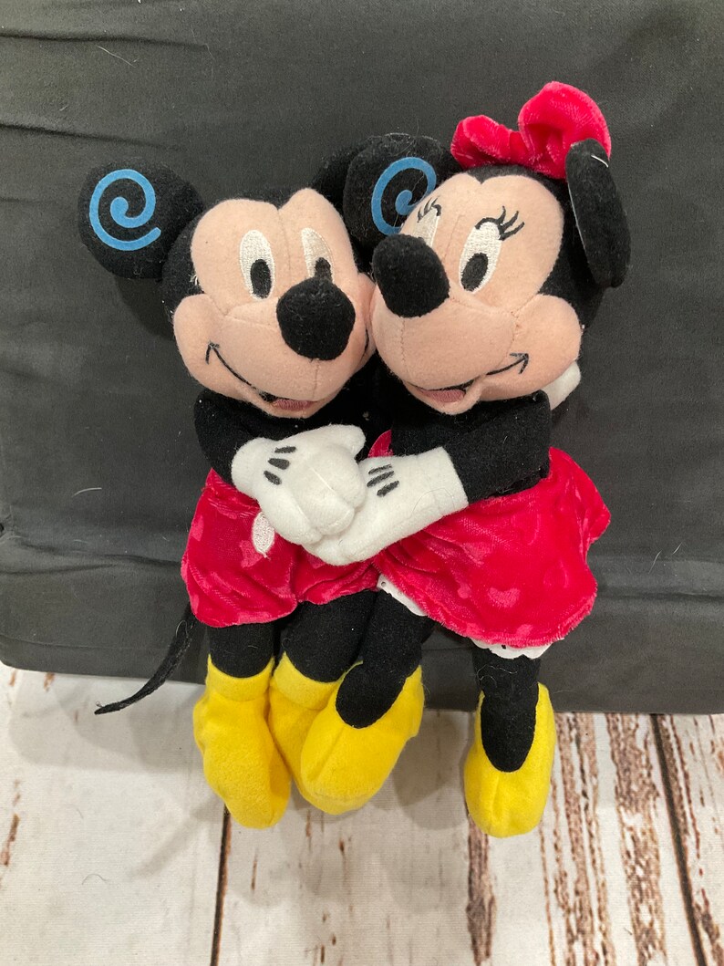 Puede incluir: Una pareja de peluche de Mickey Mouse y Minnie Mouse tomados de la mano. Mickey lleva pantalones cortos rojos y zapatos amarillos. Minnie lleva un vestido rojo con lunares blancos y zapatos amarillos. Ambos est&aacute;n sonriendo.