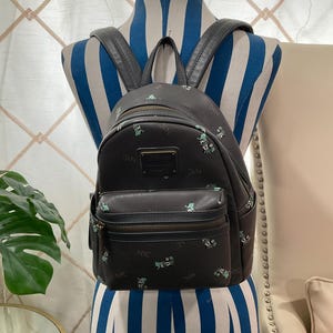 May include: Black backpack with a small front pocket and top handle. The backpack has a repeating pattern of small green and white illustrations and the letters "NYC". The backpack is displayed on a blue and white striped mannequin.