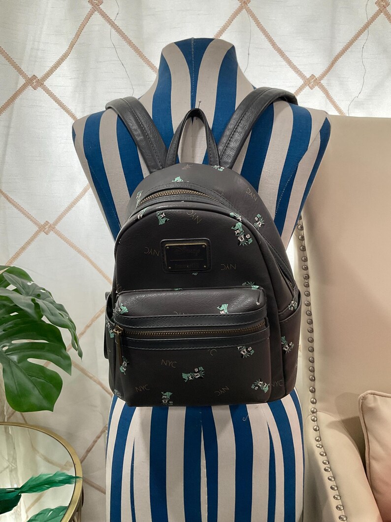 May include: Black backpack with a small front pocket and top handle. The backpack has a repeating pattern of small green and white illustrations and the letters "NYC". The backpack is displayed on a blue and white striped mannequin.
