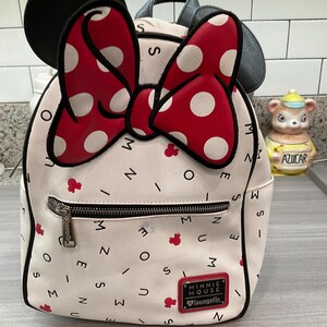 May include: White Minnie Mouse mini backpack with a red polka dot bow and black ears. The bag has a front zipper pocket and a small red Minnie Mouse logo. The bag is decorated with red Minnie Mouse icons and the words "Minnie Mouse" and "Loungefly".