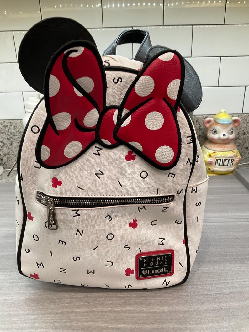 May include: White Minnie Mouse mini backpack with a red polka dot bow and black ears. The bag has a front zipper pocket and a small red Minnie Mouse logo. The bag is decorated with red Minnie Mouse icons and the words "Minnie Mouse" and "Loungefly".