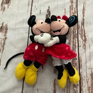 Puede incluir: Un par de peluches de Mickey Mouse y Minnie Mouse. Mickey lleva pantalones cortos rojos con lunares blancos y zapatos amarillos. Minnie lleva una falda roja con lunares blancos y zapatos amarillos. Se est&aacute;n abrazando.