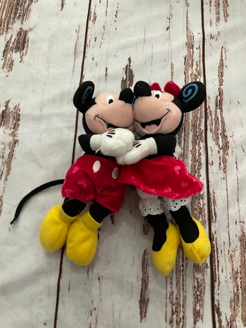 Puede incluir: Un par de peluches de Mickey Mouse y Minnie Mouse. Mickey lleva pantalones cortos rojos con lunares blancos y zapatos amarillos. Minnie lleva una falda roja con lunares blancos y zapatos amarillos. Se est&aacute;n abrazando.