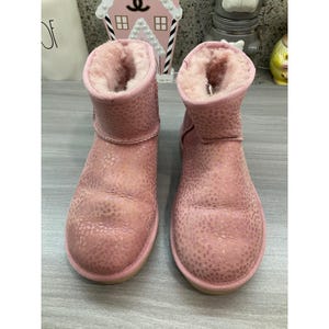May include: A pair of ankle-height boots in a soft pink hue, featuring a subtle metallic animal print pattern across the exterior. The boots are lined with a thick, plush light pink shearling material that extends to the top collar edge.