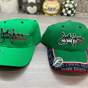 May include: Two green baseball caps with the words "John Force Racing" in black and white script. One cap has a flame design on the side, and the other has a patterned brim with the "John Force Racing" logo.