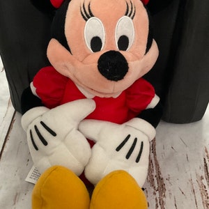 May include: A plush Minnie Mouse doll. The doll has a red dress, a red bow with white polka dots, black ears, and yellow shoes. The doll's hands are white with black gloves. The doll is sitting on a wooden surface.